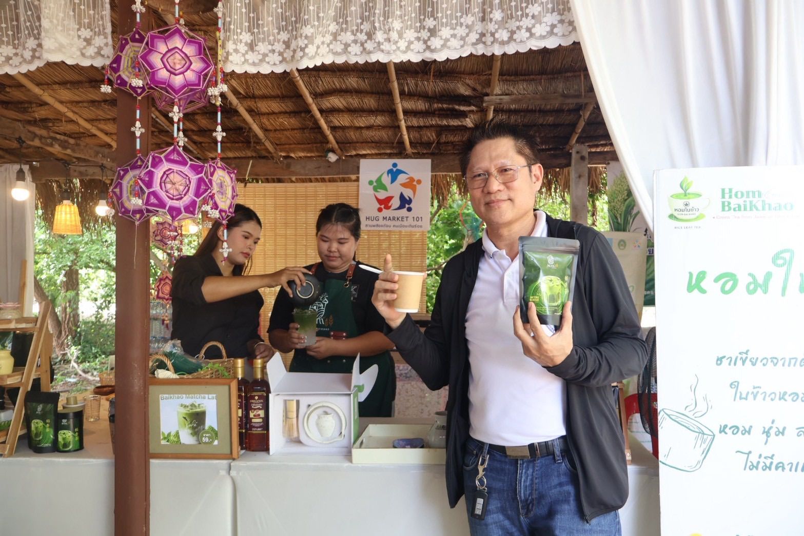 A man holds a green product package and gives a thumbs-up at an outdoor market stall.
