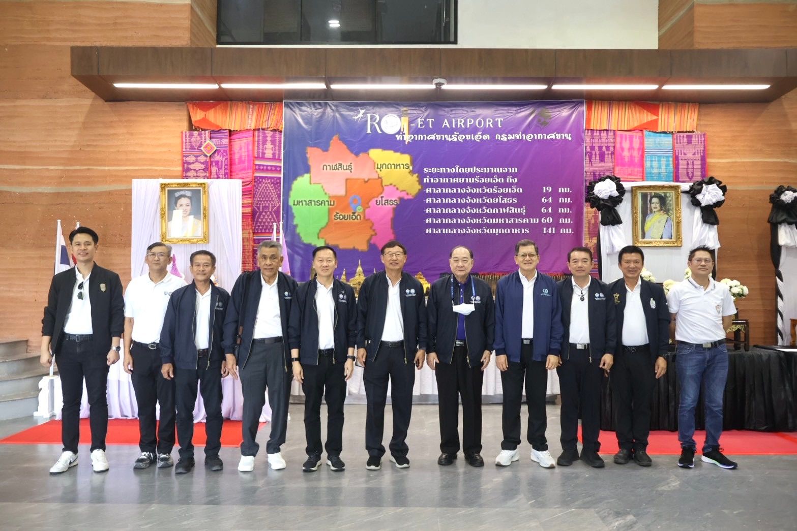 A group of formally dressed men stands smiling in front of a banner at an event.