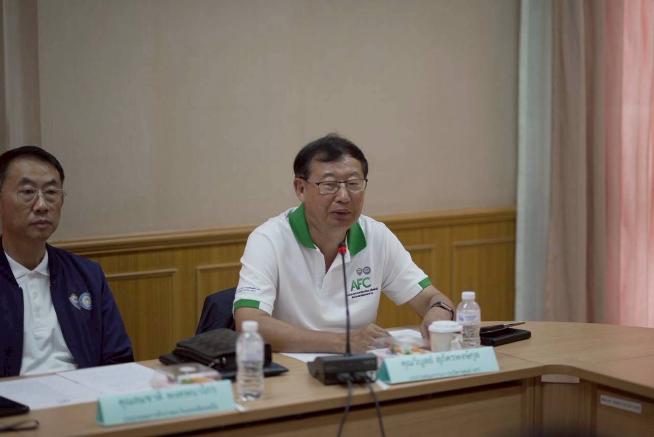 Man in white polo shirt speaks into microphone at a conference table beside another man.