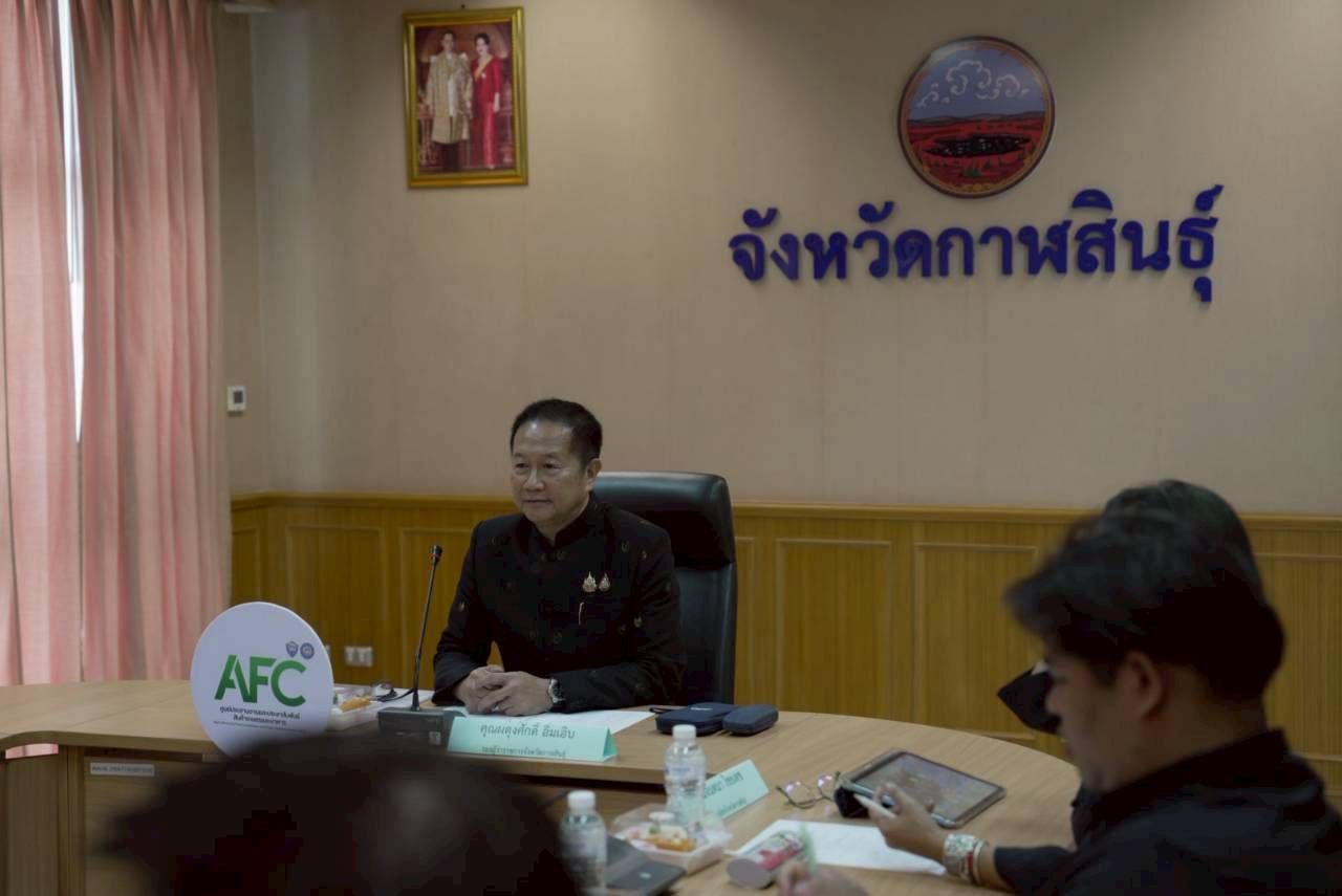 Man in dark shirt sits at conference table with Thai text on the wall.
