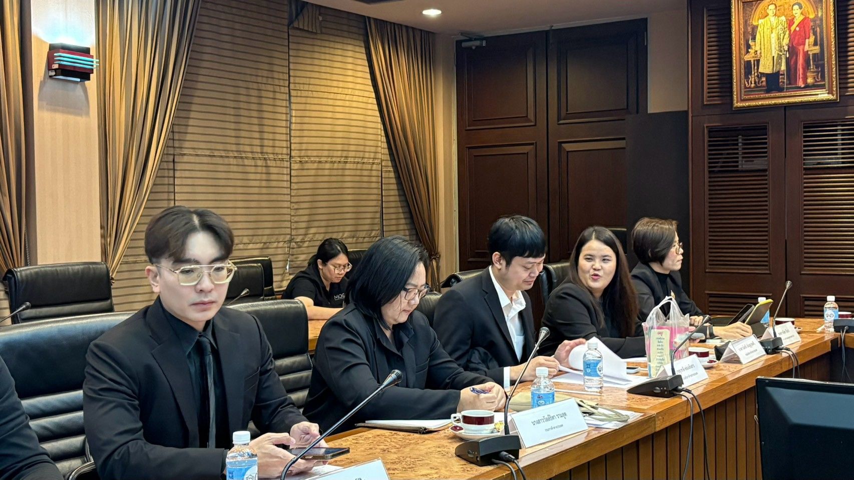 Professionals in dark suits attend a formal meeting in a wood-paneled conference room.
