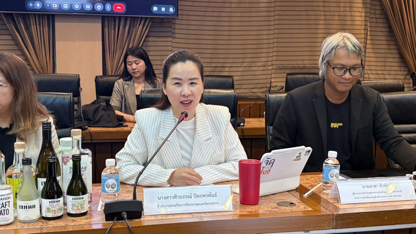 A woman in a white blazer speaks into a microphone at a formal conference table.