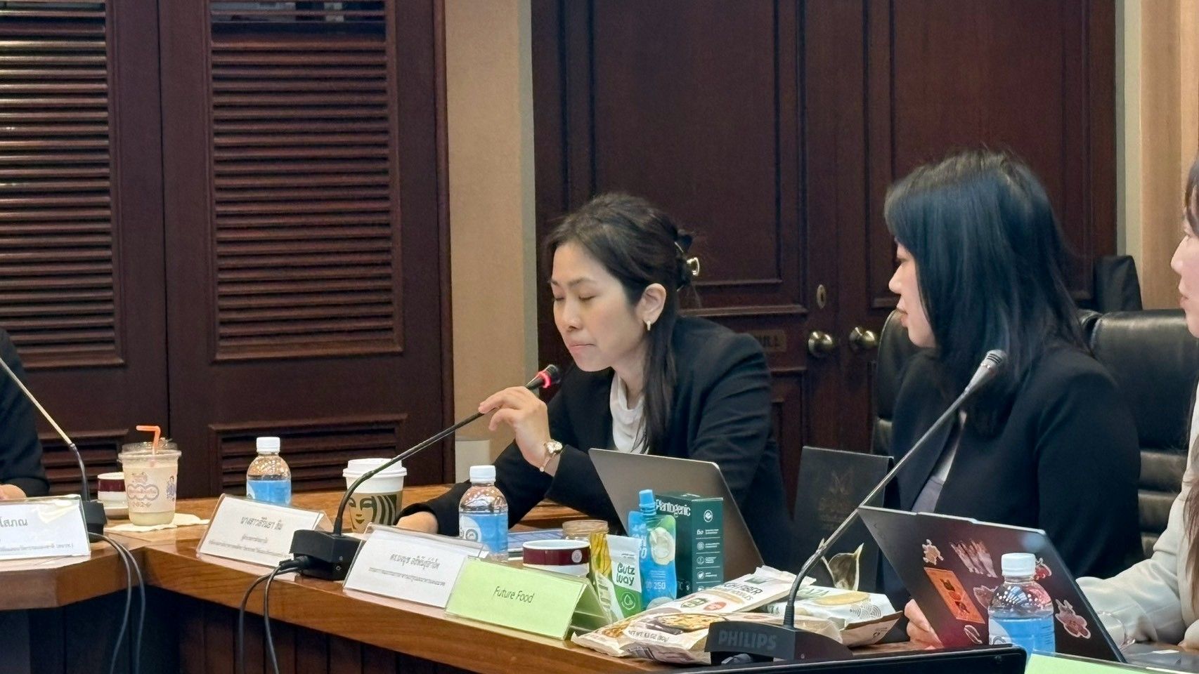 Woman in dark suit speaks into a microphone at a conference table with other attendees.