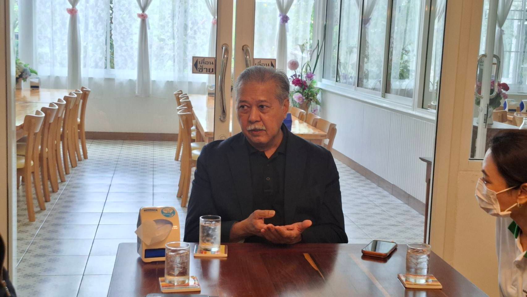 Elderly man in black shirt gestures while speaking at a wooden table indoors.