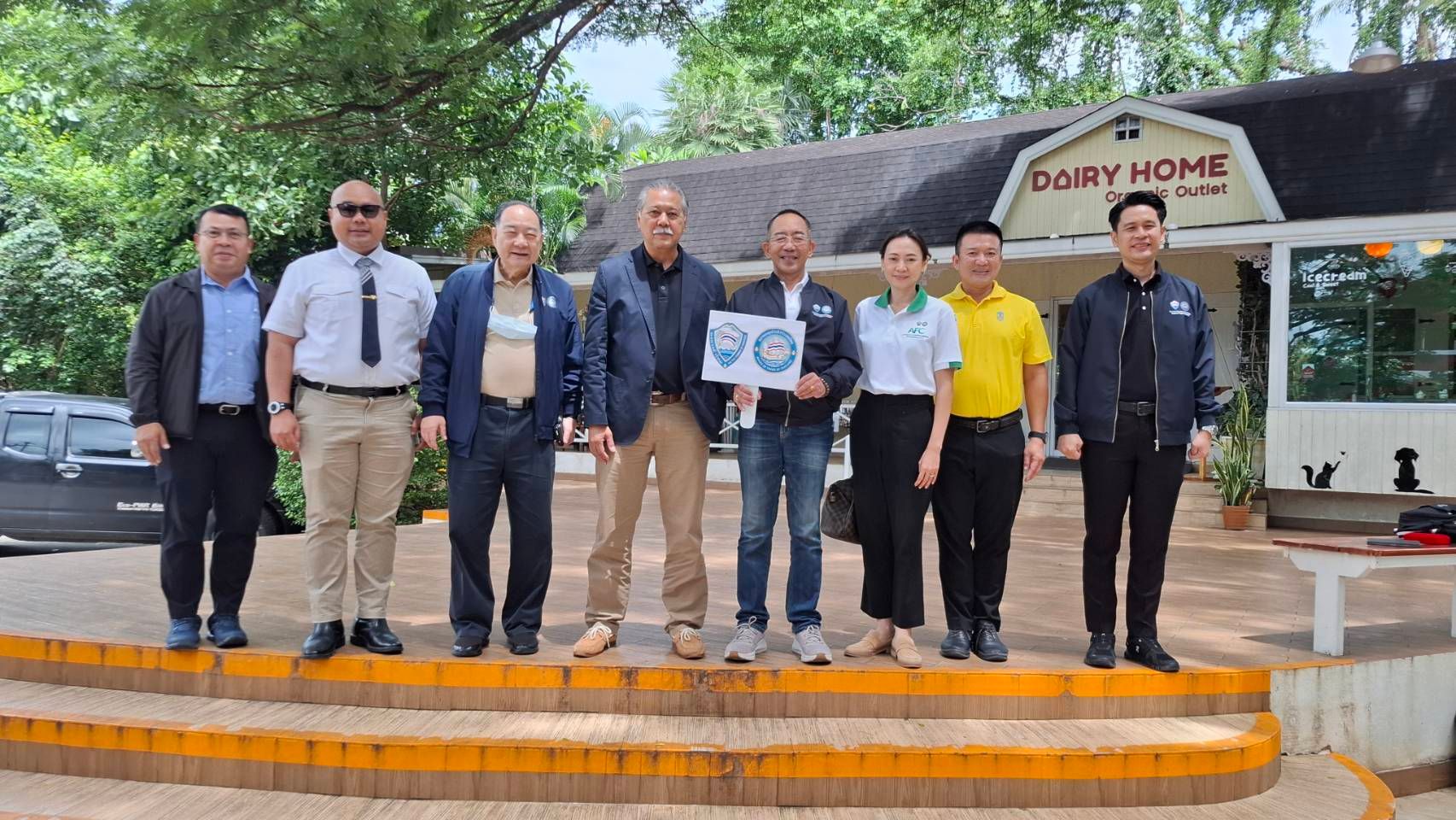 Eight people, some in uniform, stand on steps holding an award in front of Dairy Home.