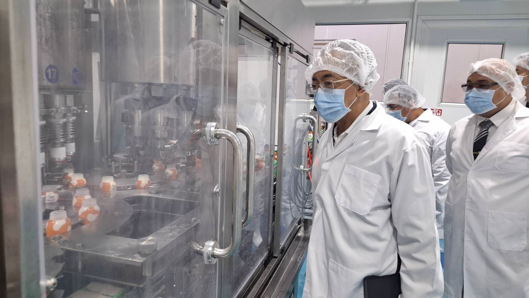 Inspector in lab coat and mask observes an automated bottling production line in a sterile facility.
