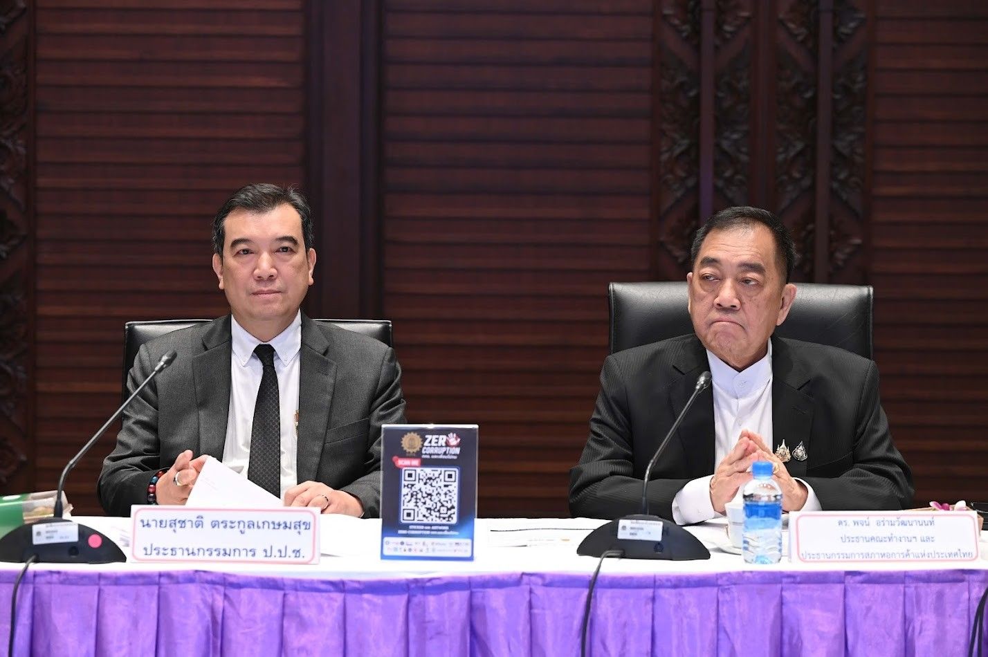 Two formally dressed men sit at a conference table with a QR code stand visible.