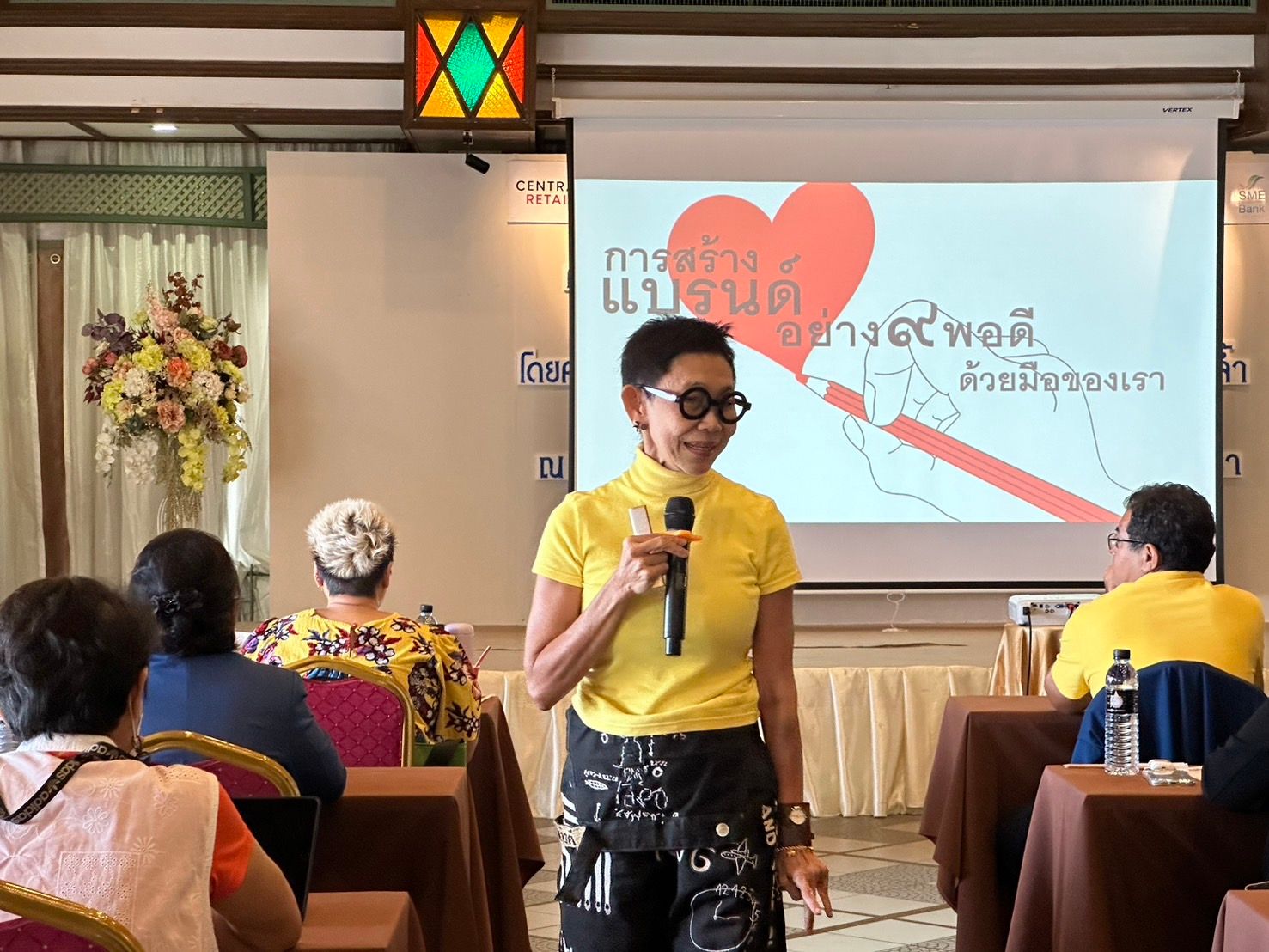 Woman in yellow shirt speaking into microphone during a presentation to an audience.