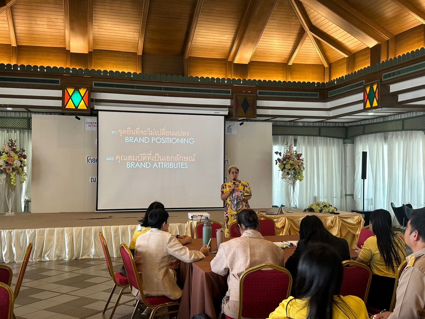 A speaker presents to an audience seated at tables in a large conference room.