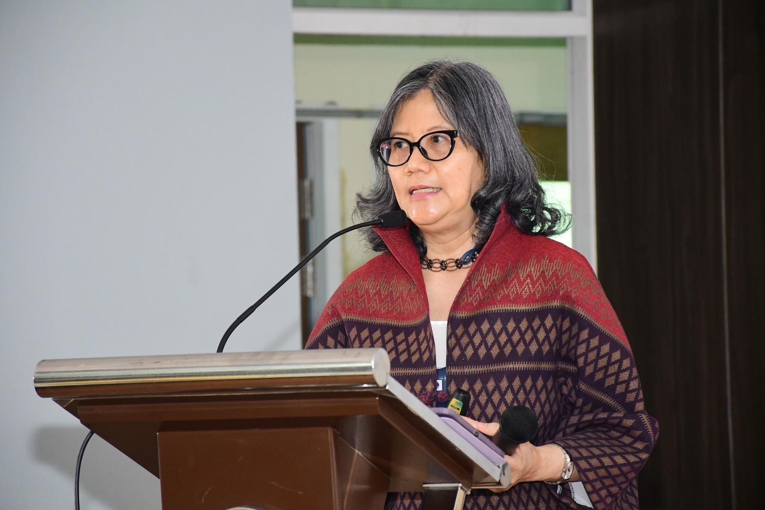 A woman with gray hair and glasses speaks at a podium wearing a patterned shawl.