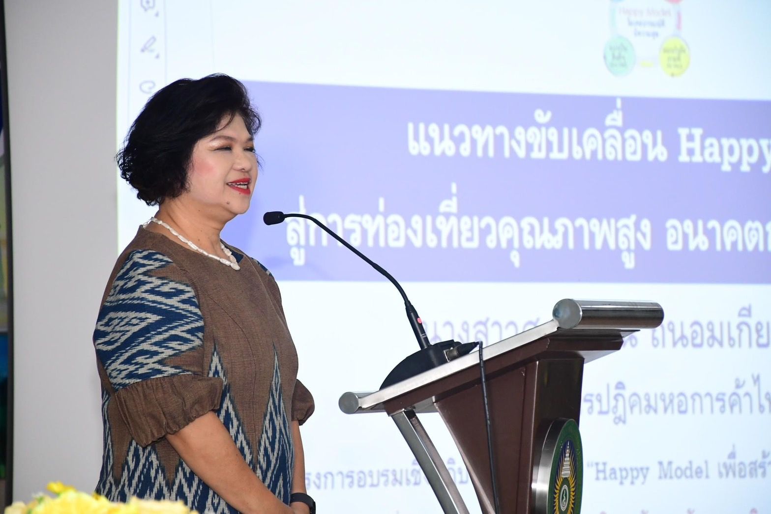 A woman with short dark hair speaks at a podium during a presentation on Happy Tourism.