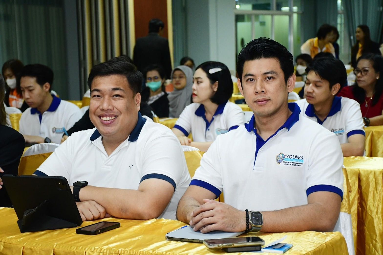 Two men in white and blue polo shirts sitting at a conference table, looking forward.