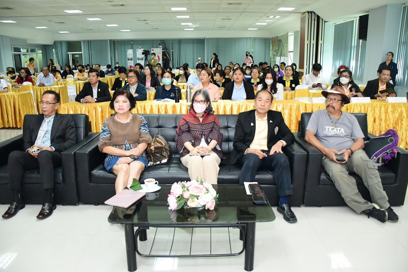 Five attendees on a black couch at a conference with an audience in the background.