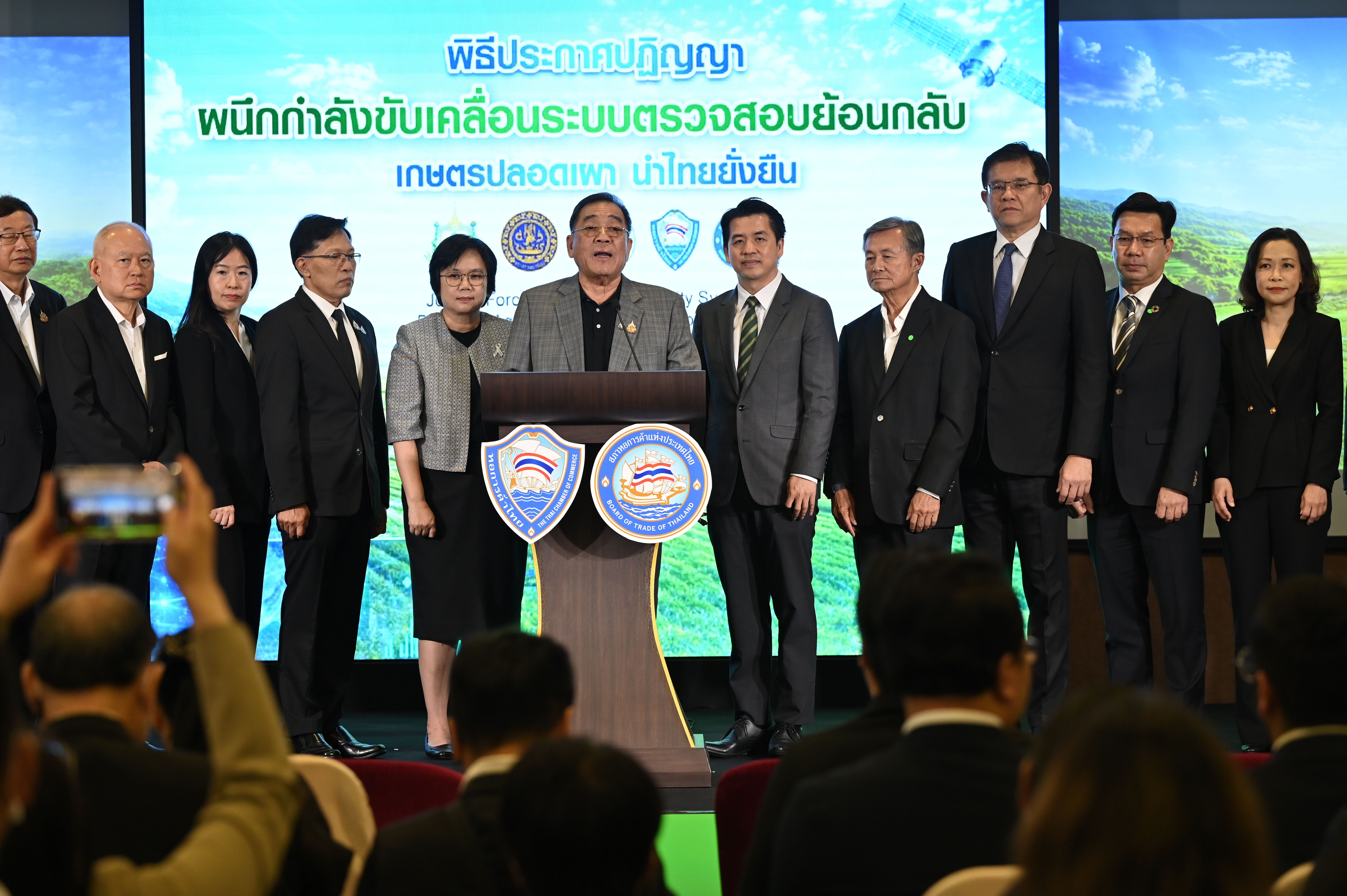 Official speaks at podium, flanked by colleagues, under screen with Thai text.