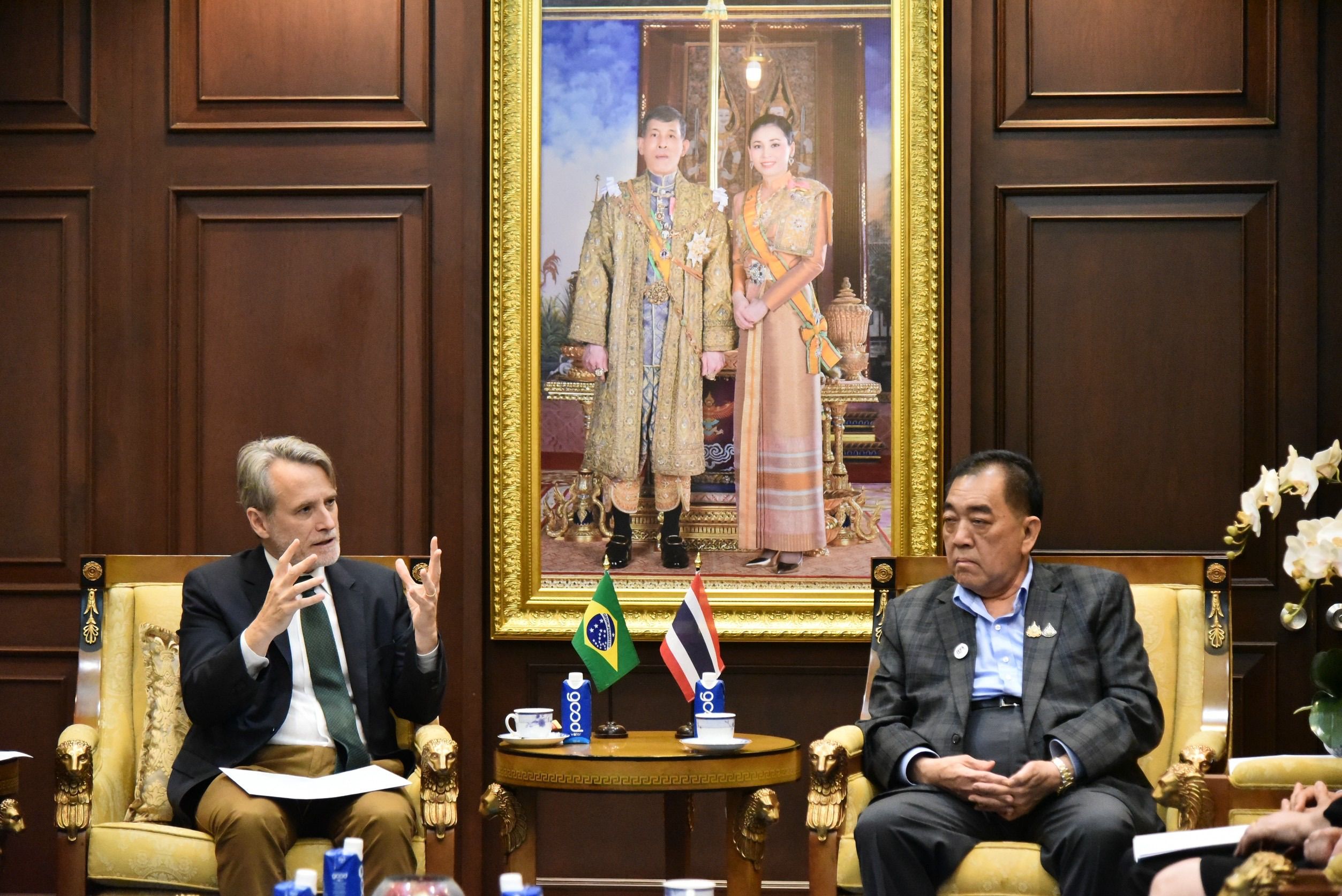 A diplomatic meeting between two officials with national flags and a royal portrait.