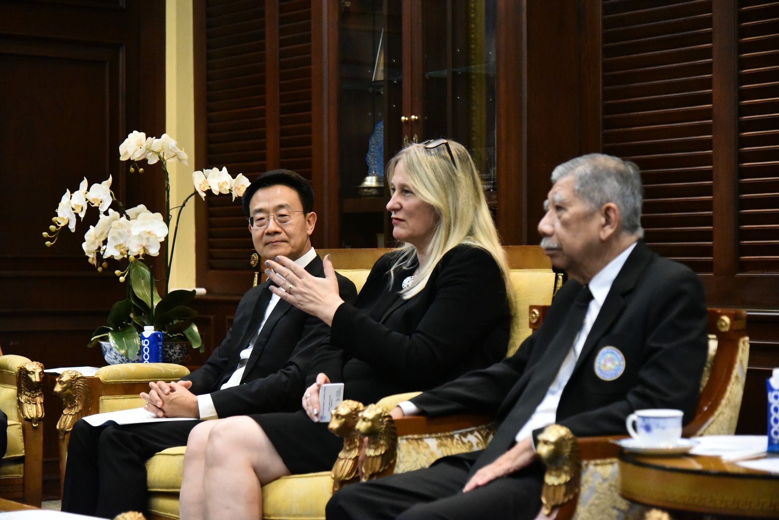 Woman speaking to two men in a formal meeting, seated in ornate chairs.