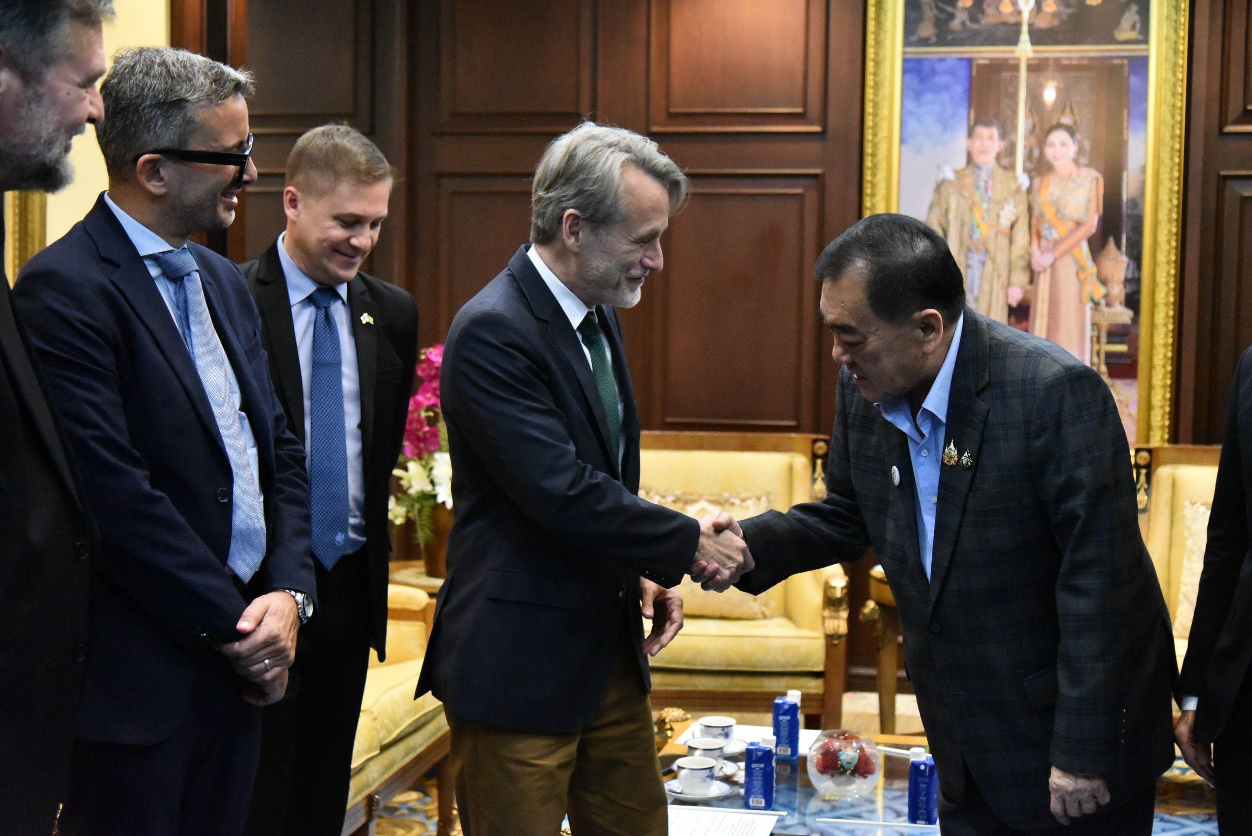 Two men in suits shake hands warmly in a formal meeting room, observed by other officials.