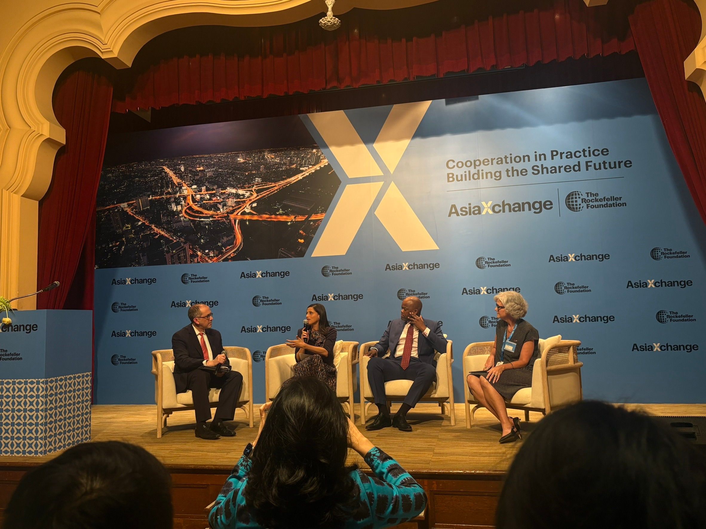 Four speakers present a panel discussion on stage with a blue banner featuring an 'X' logo.