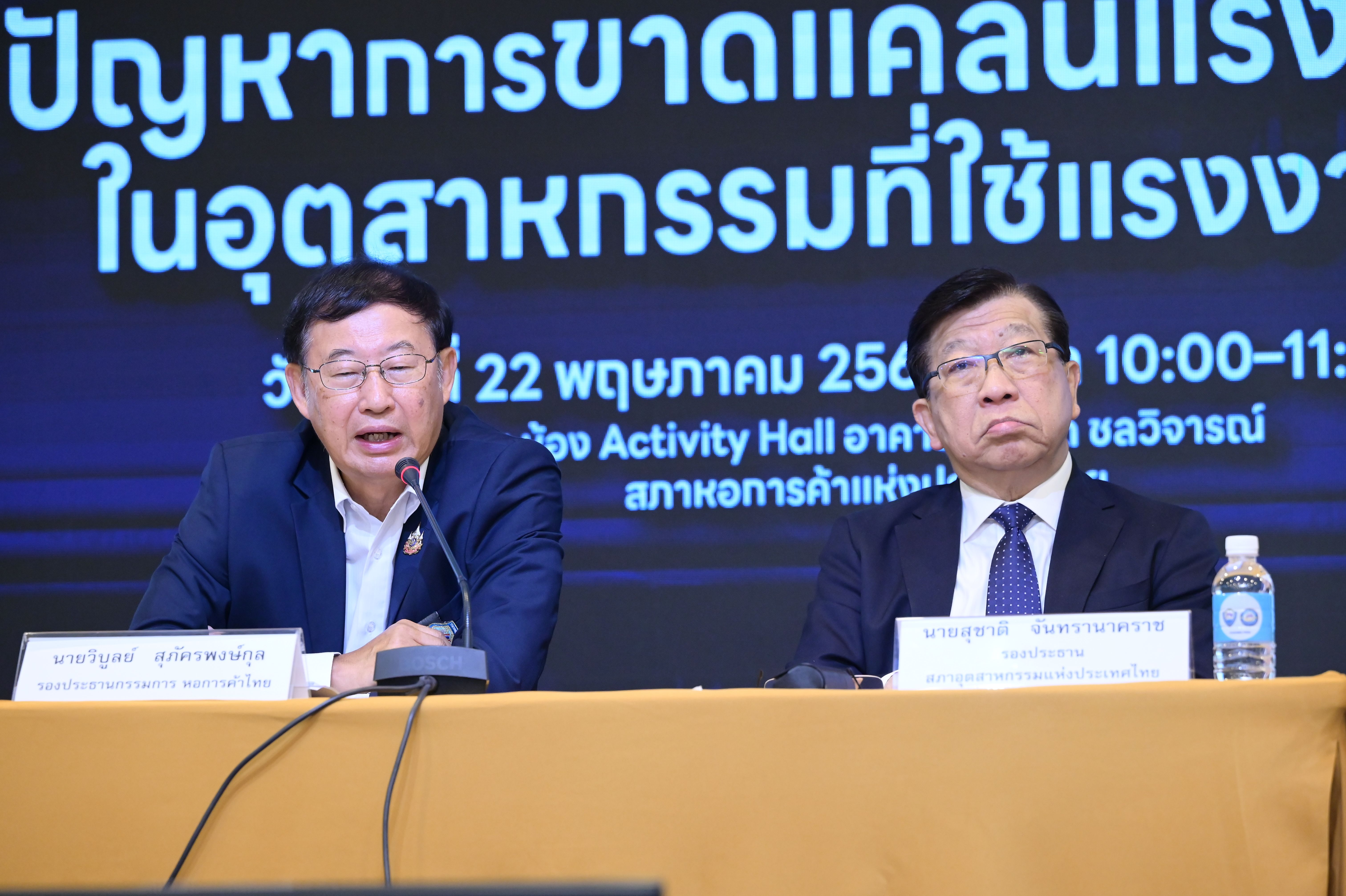 Two executives speak at a conference with Thai text on a screen behind them.