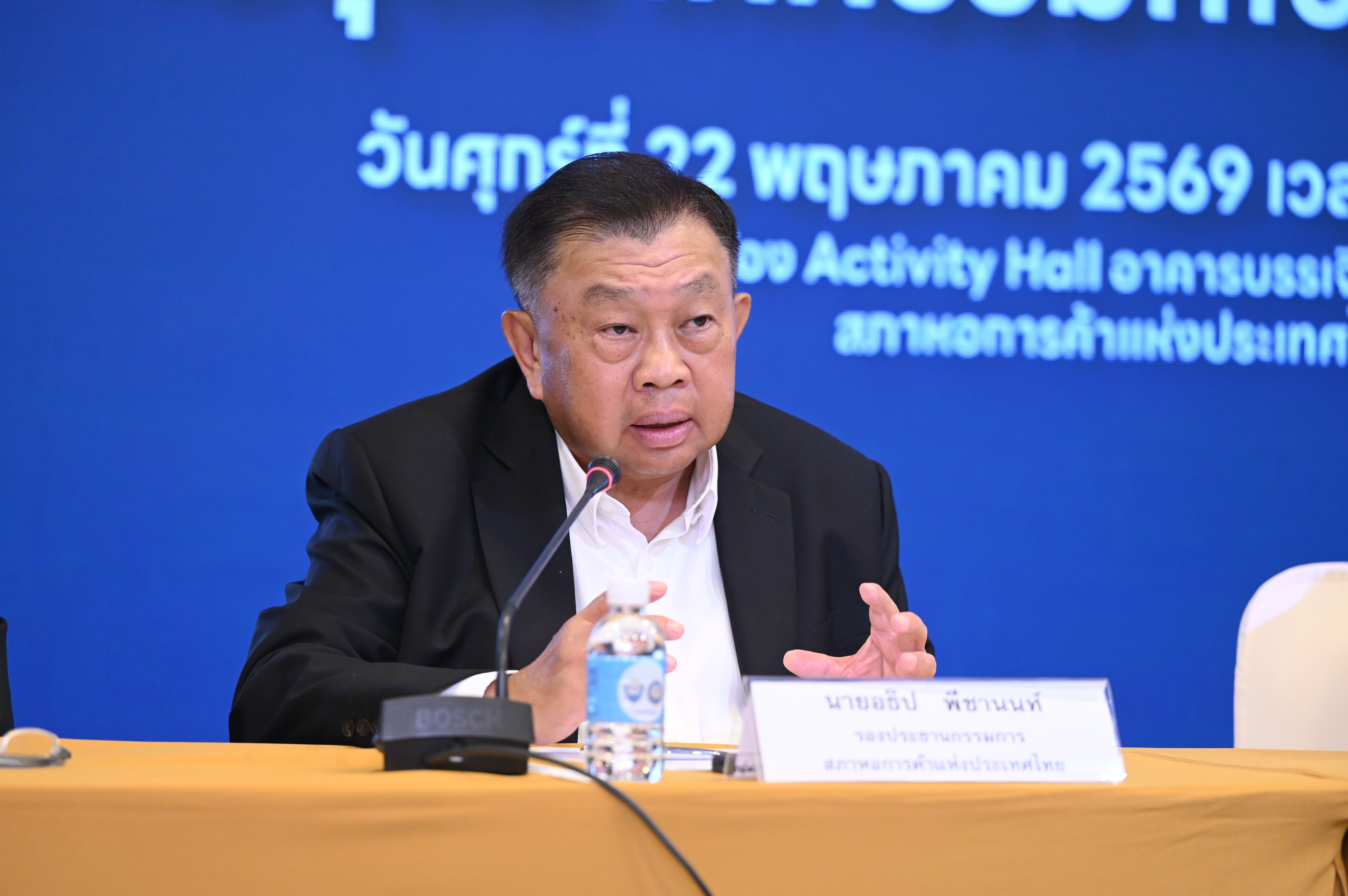 Older Asian man in a suit speaks into a microphone at a conference table.