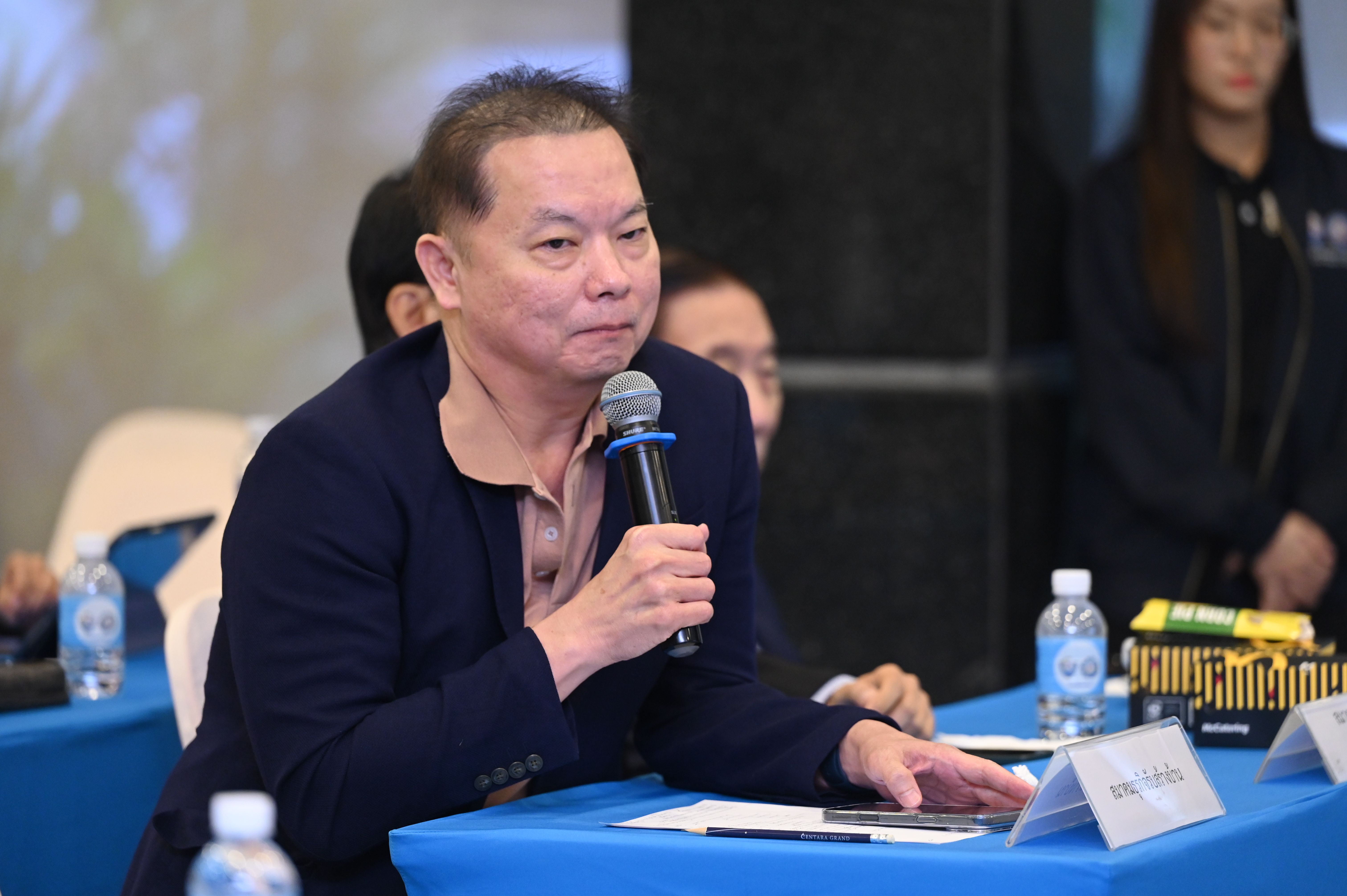 Elderly Asian man speaking intently into a microphone at a formal event.