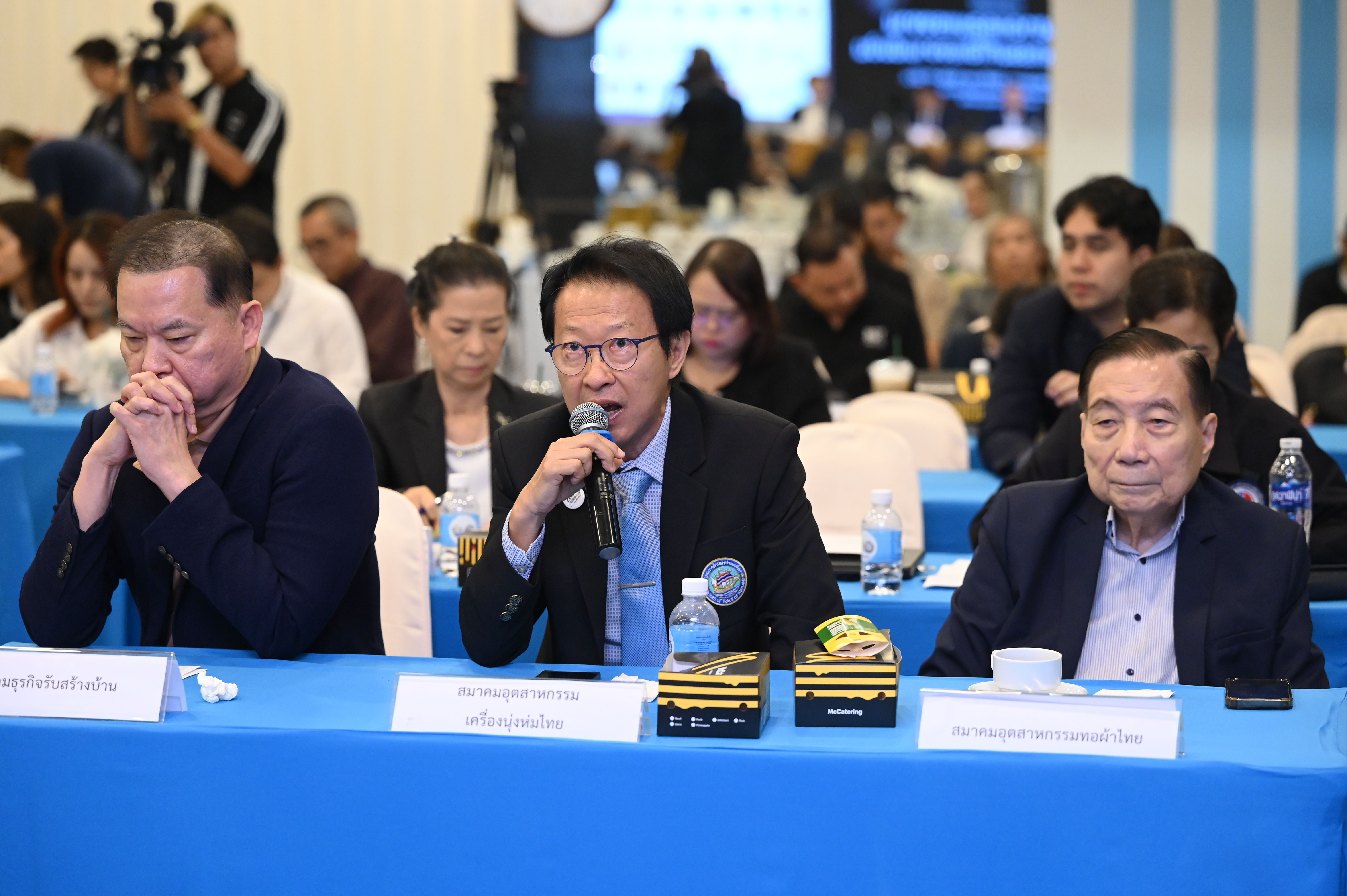 A man speaks into a microphone at a table, flanked by two other men.