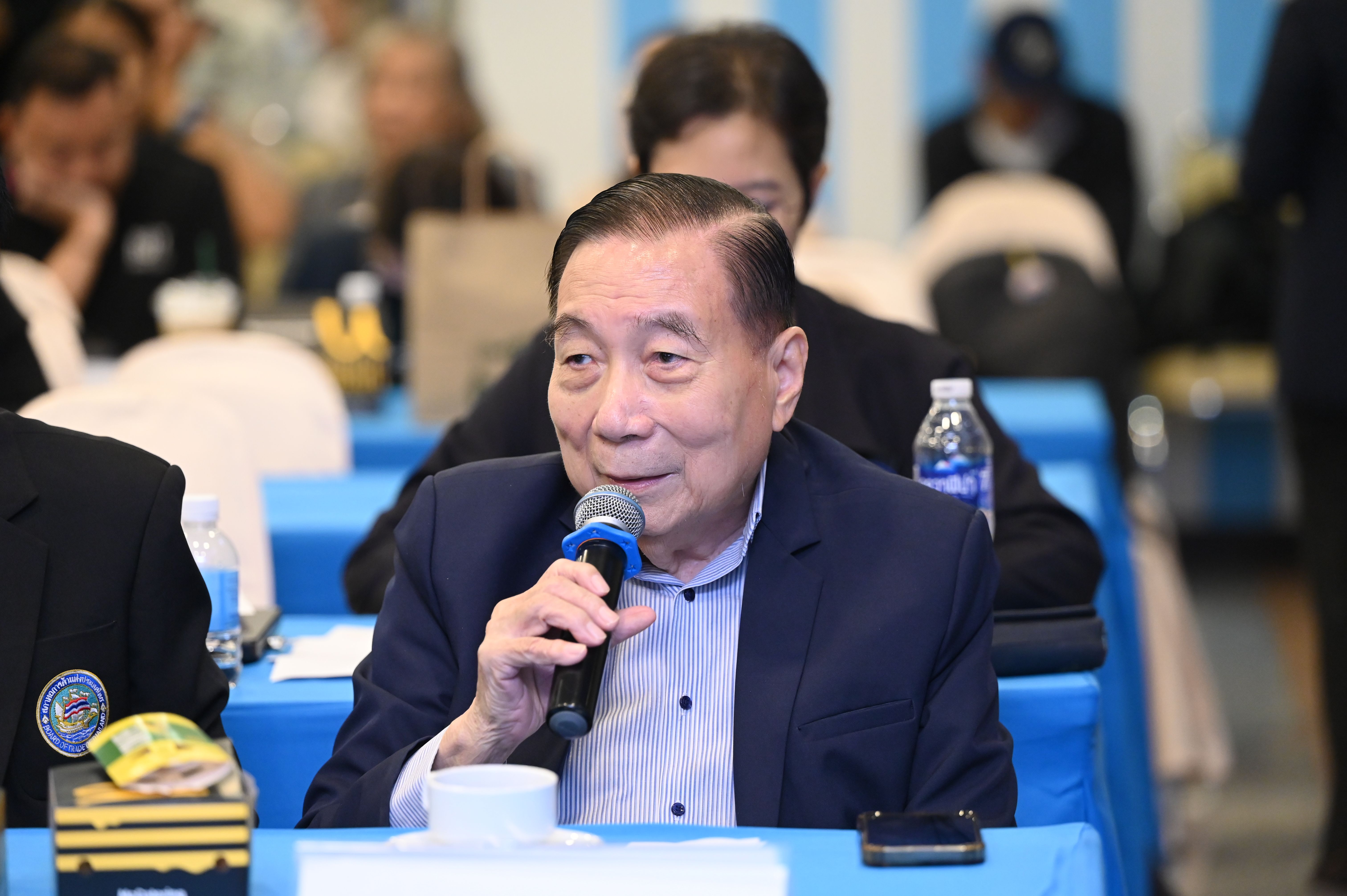 Elderly Asian man speaks into a microphone while seated at a formal event.