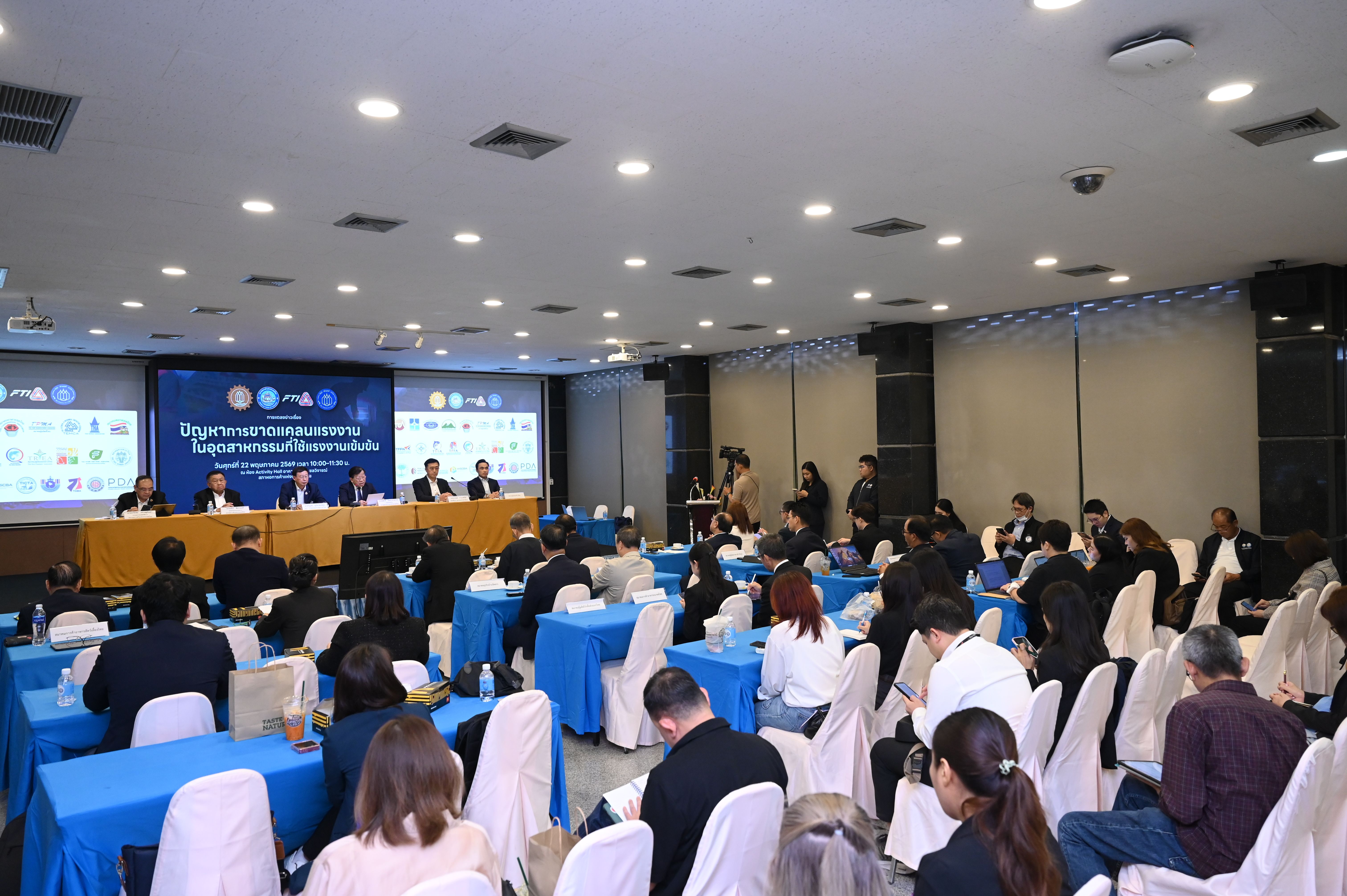 Audience seated in a large conference room, watching presenters on a stage with screens.