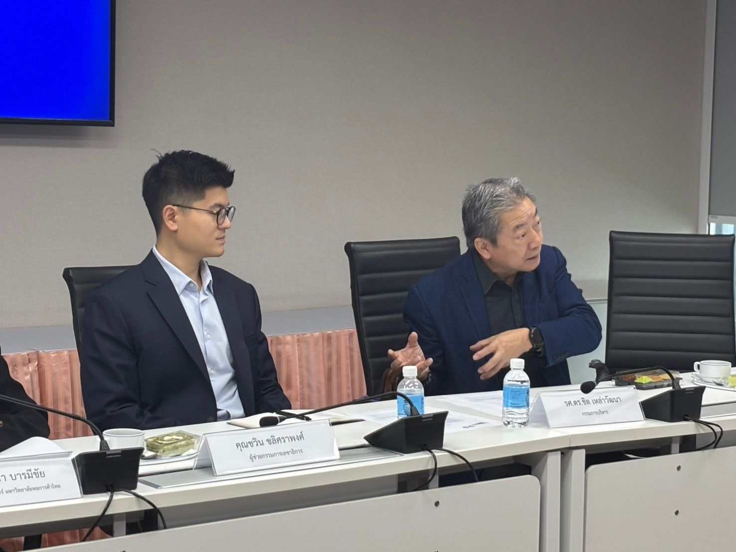 Two men, one younger and one older, discuss at a conference table.
