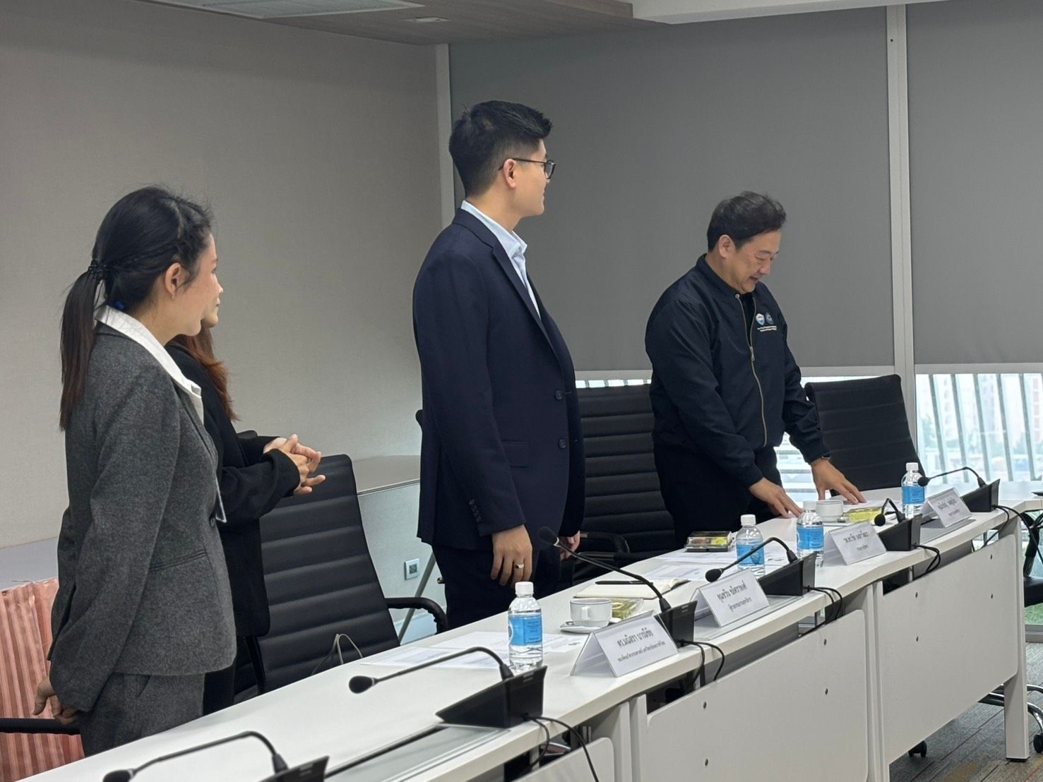 Three professionals stand around a conference table with name placards, water, and microphones.