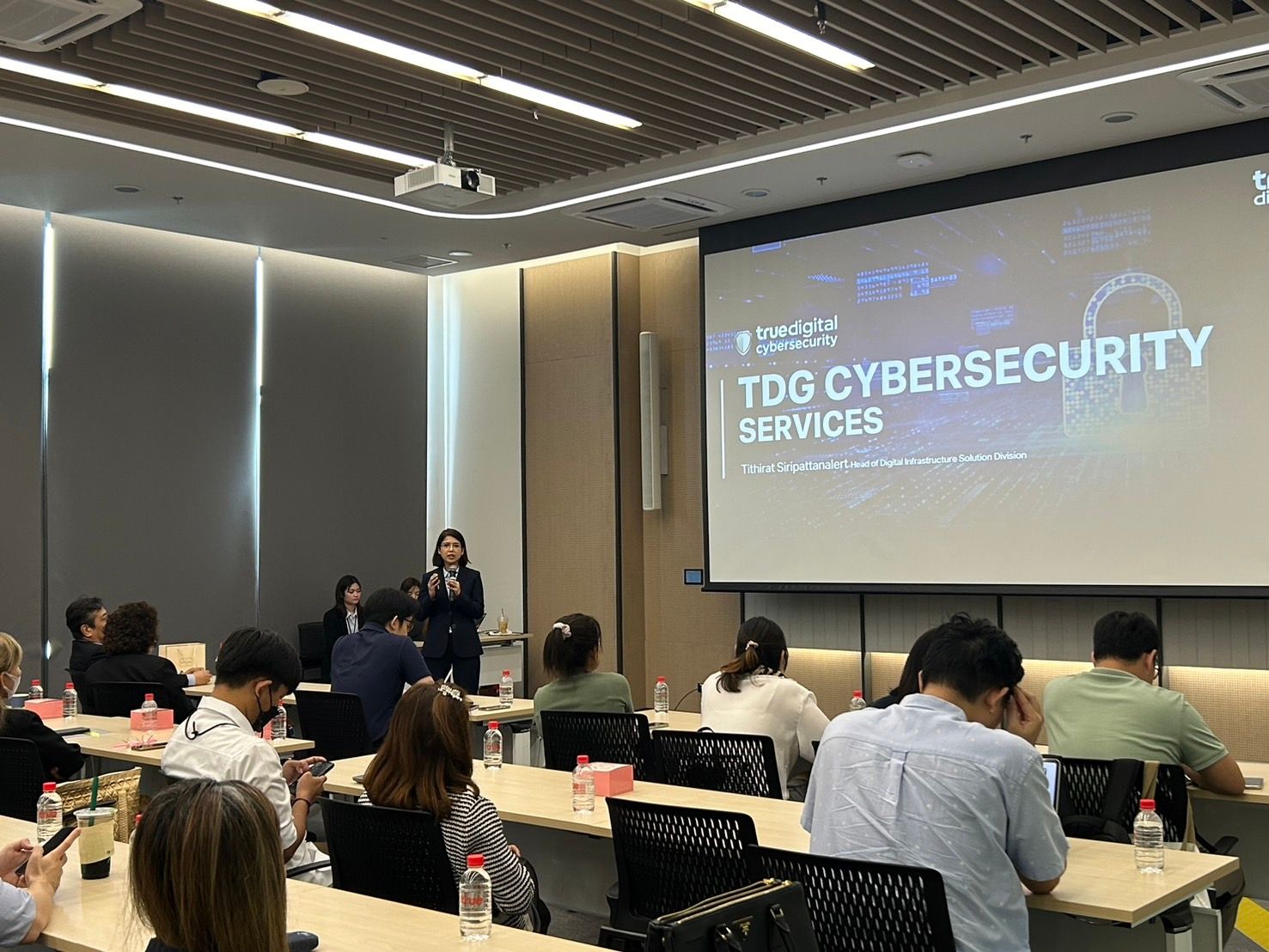 A speaker presents 'TDG Cybersecurity Services' to an attentive audience in a modern conference room.