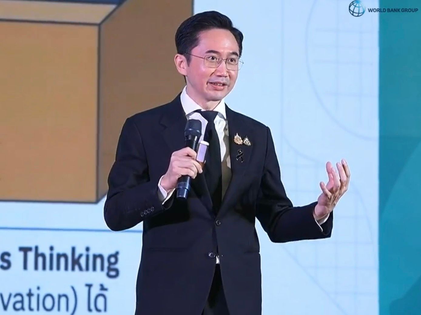 A man in a suit and glasses speaks into a microphone at a presentation.