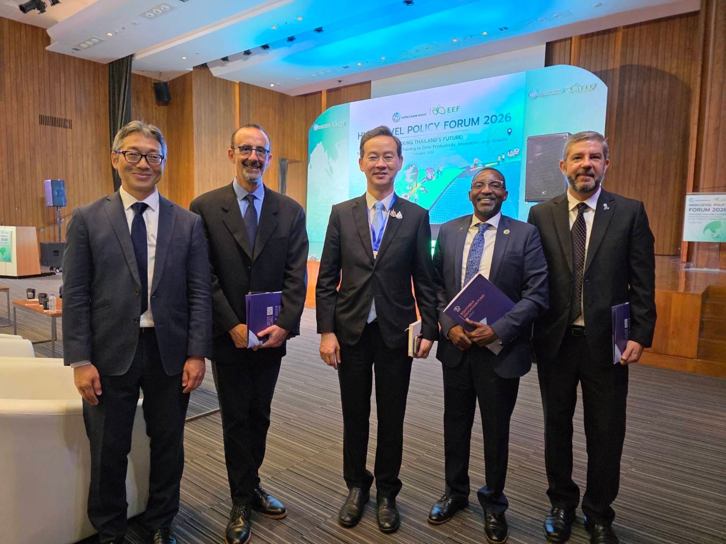 Five men in suits stand together at a professional conference, holding documents.