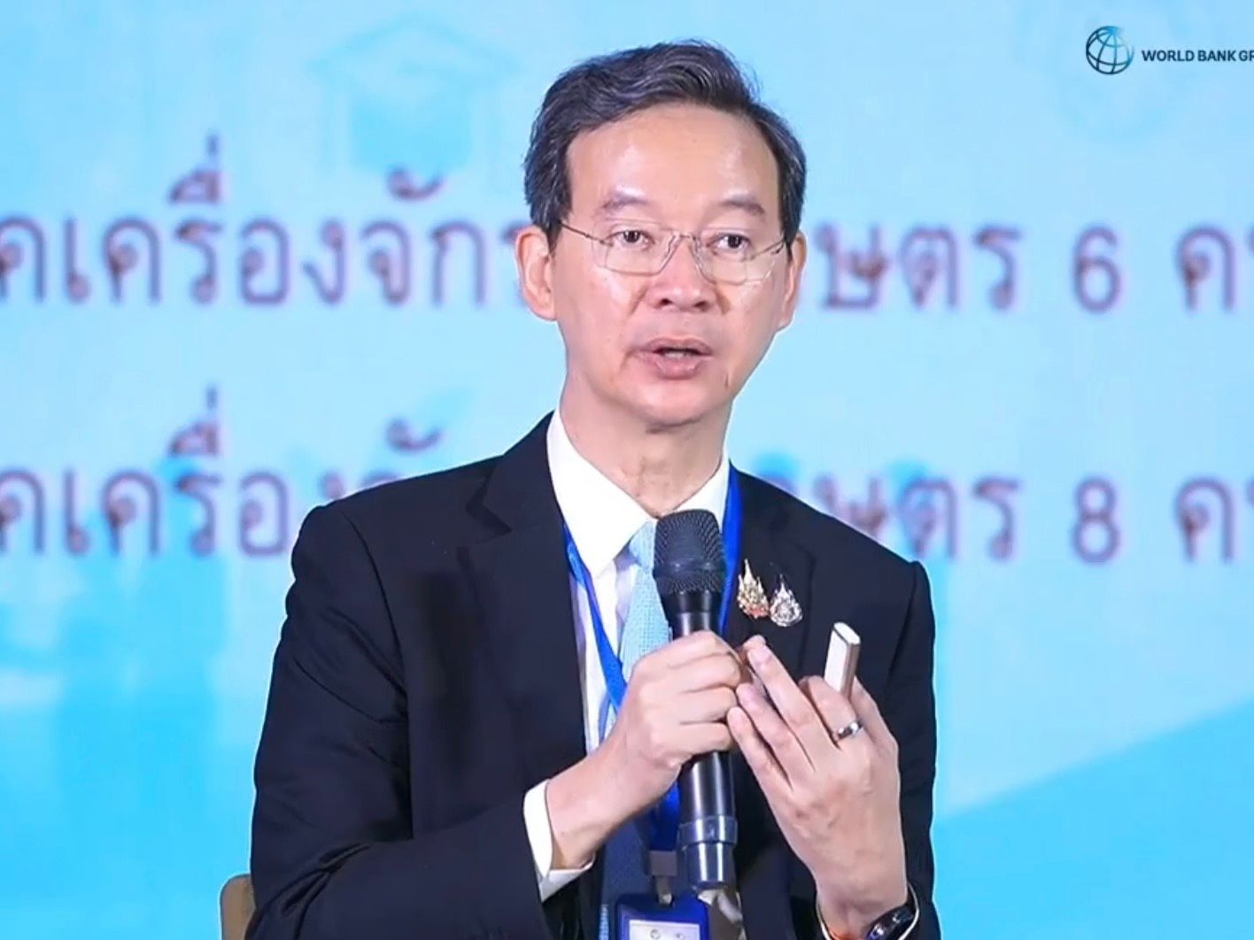 A man in a suit and glasses speaks at a podium, gesturing with his hands.
