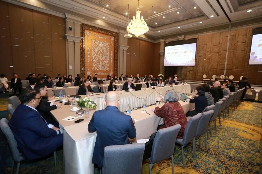 Attendees at a formal conference sit around a U-shaped table with a presentation screen.