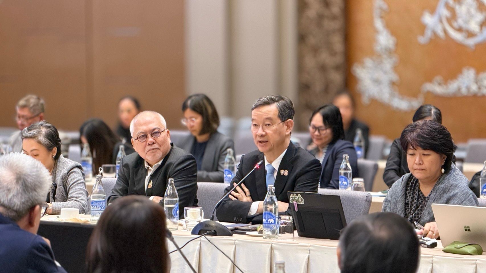 Asian man in suit speaks into microphone at a formal conference table.
