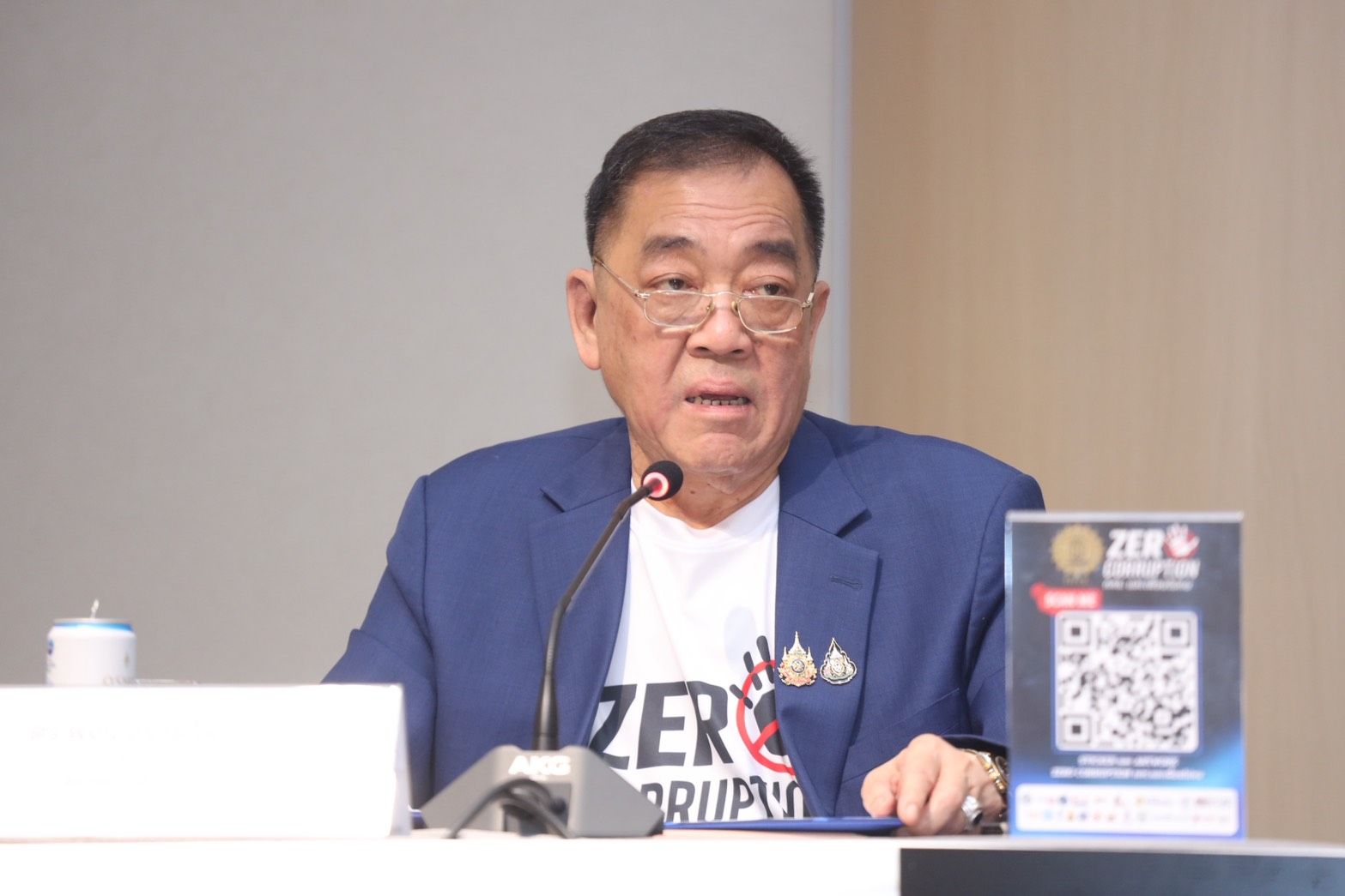 Man in blue jacket and glasses speaking at a podium with a "ZERO CORRUPTION" shirt.
