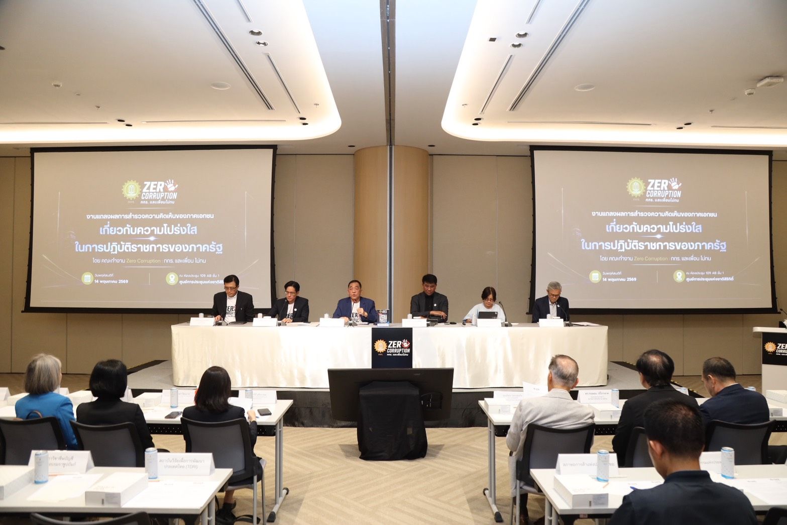 A panel of speakers addresses an audience in a conference room with large presentation screens.