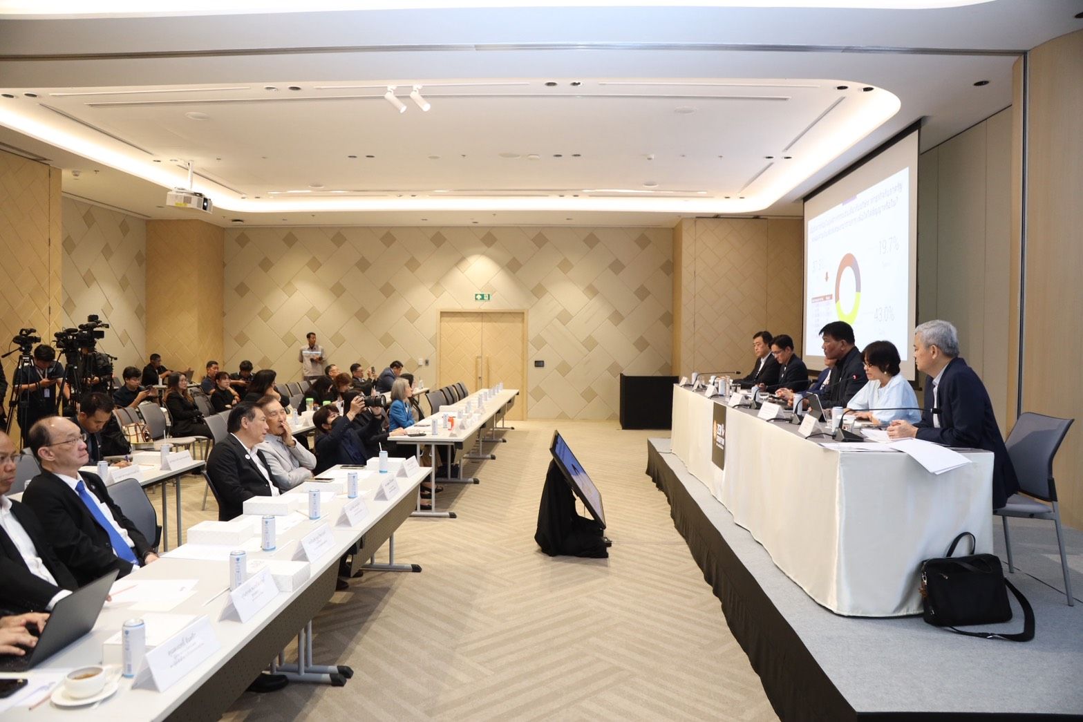 Attendees listen intently at a formal conference with a panel of speakers and projector screen.