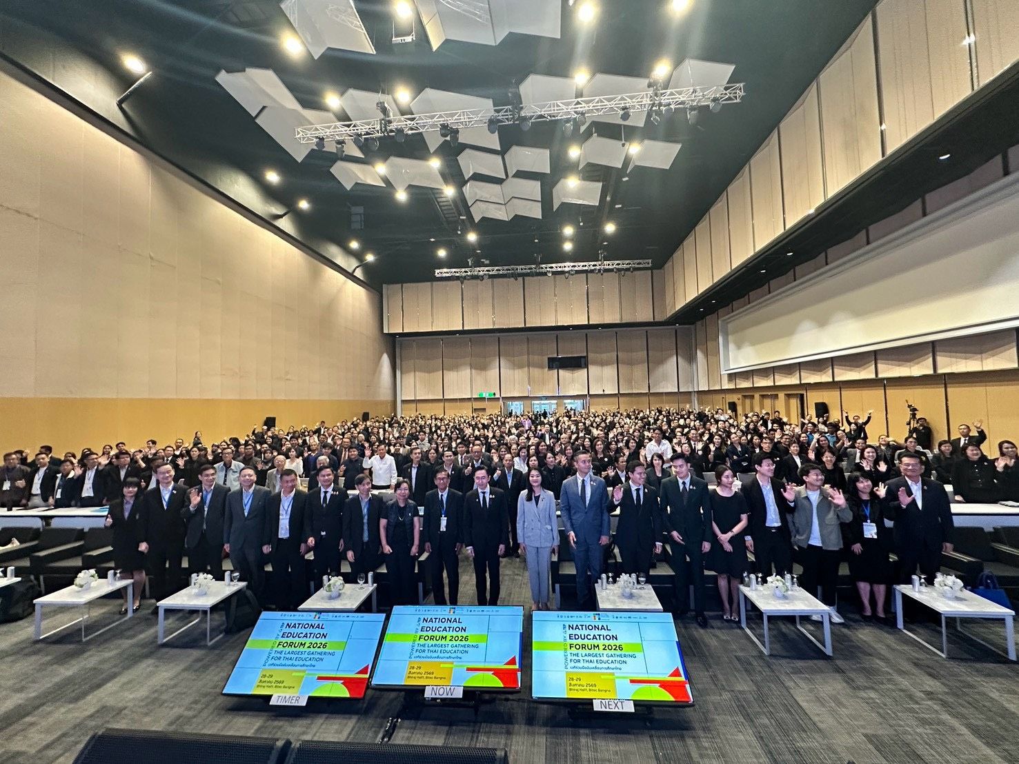 A large group of professionals stands in a spacious conference hall with three illuminated screens.