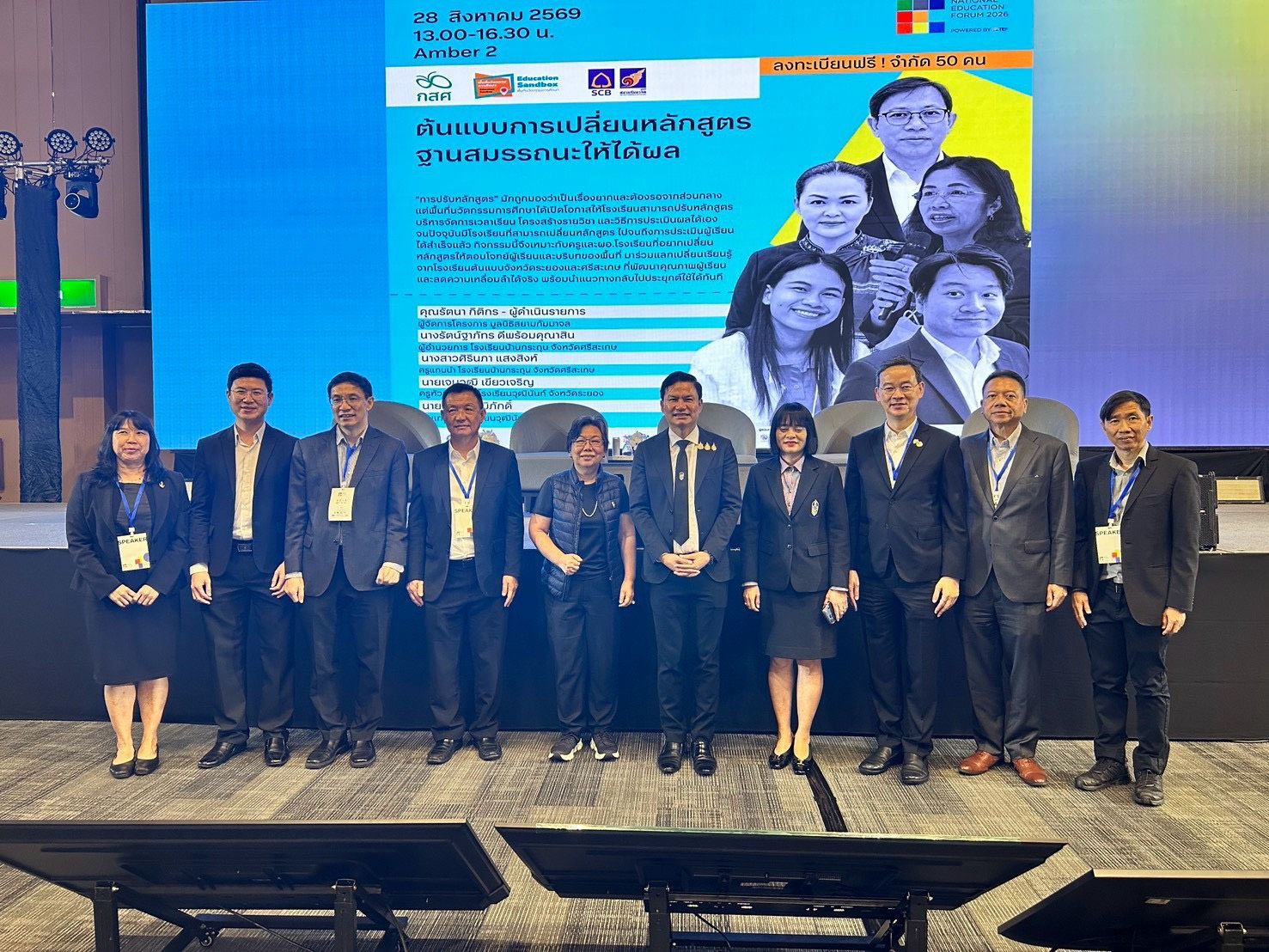 A group of professionals stands on a stage with a large screen displaying information and portraits.