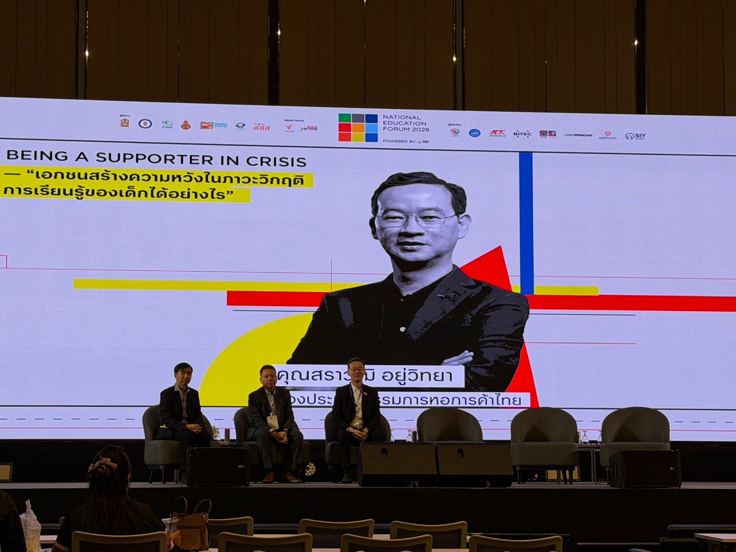 A presentation screen shows a speaker's headshot, event title, and three panelists on stage.