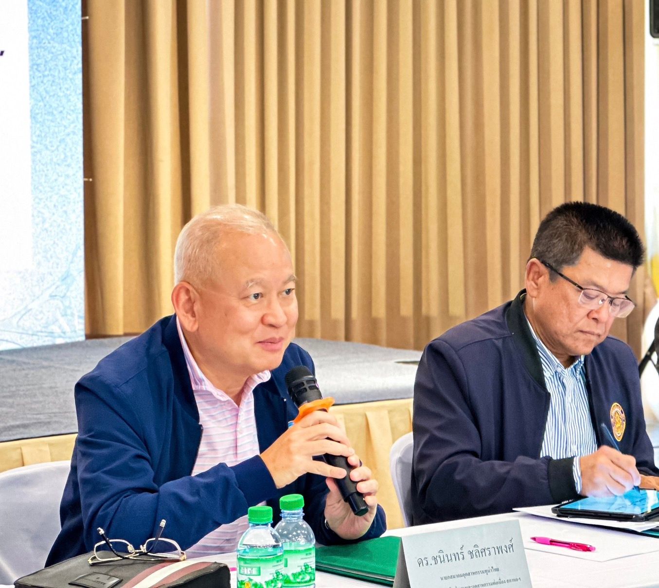 Man in blue jacket speaks into a microphone at a conference table.