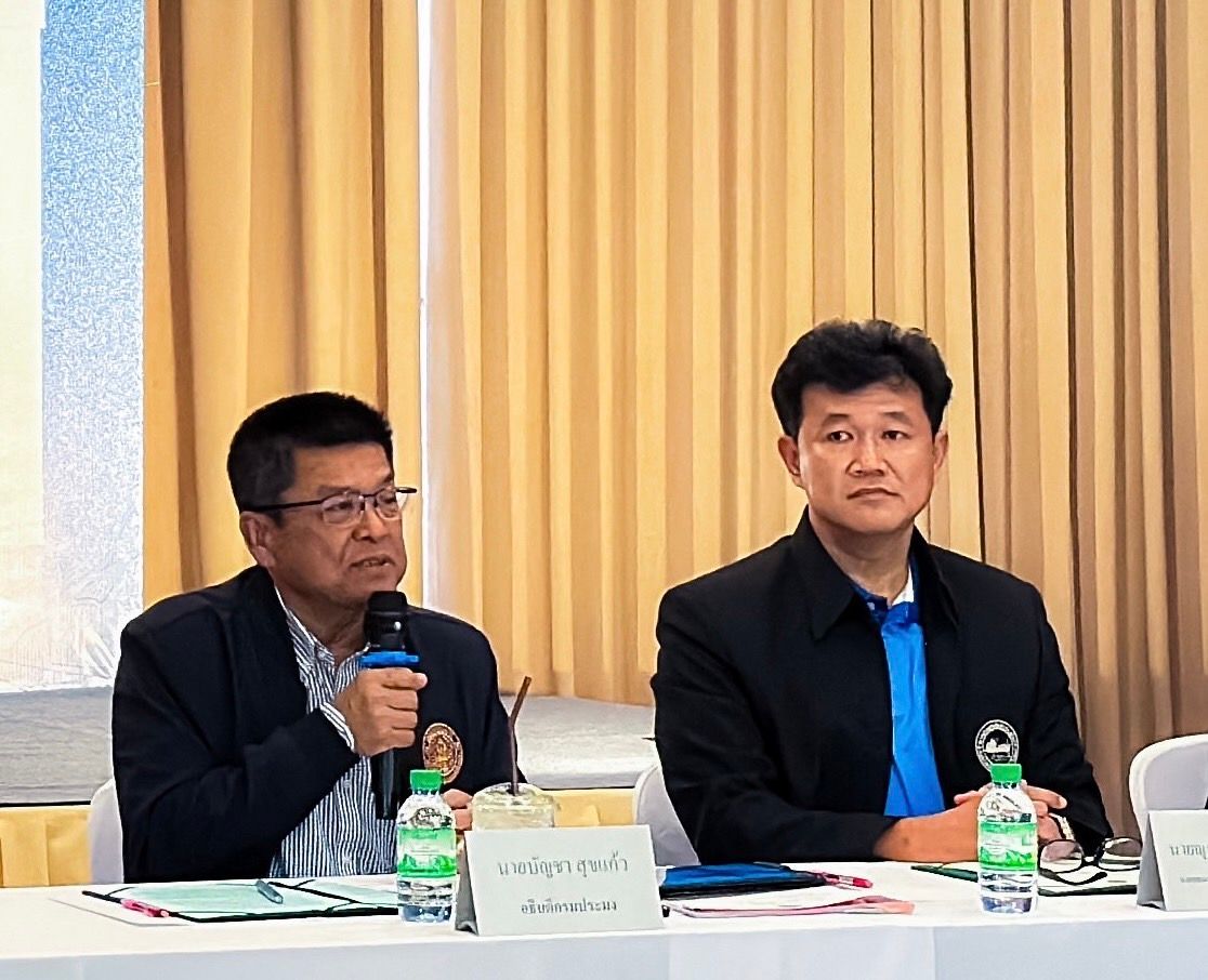 Two men at a meeting table, one speaking into a microphone, the other listening.