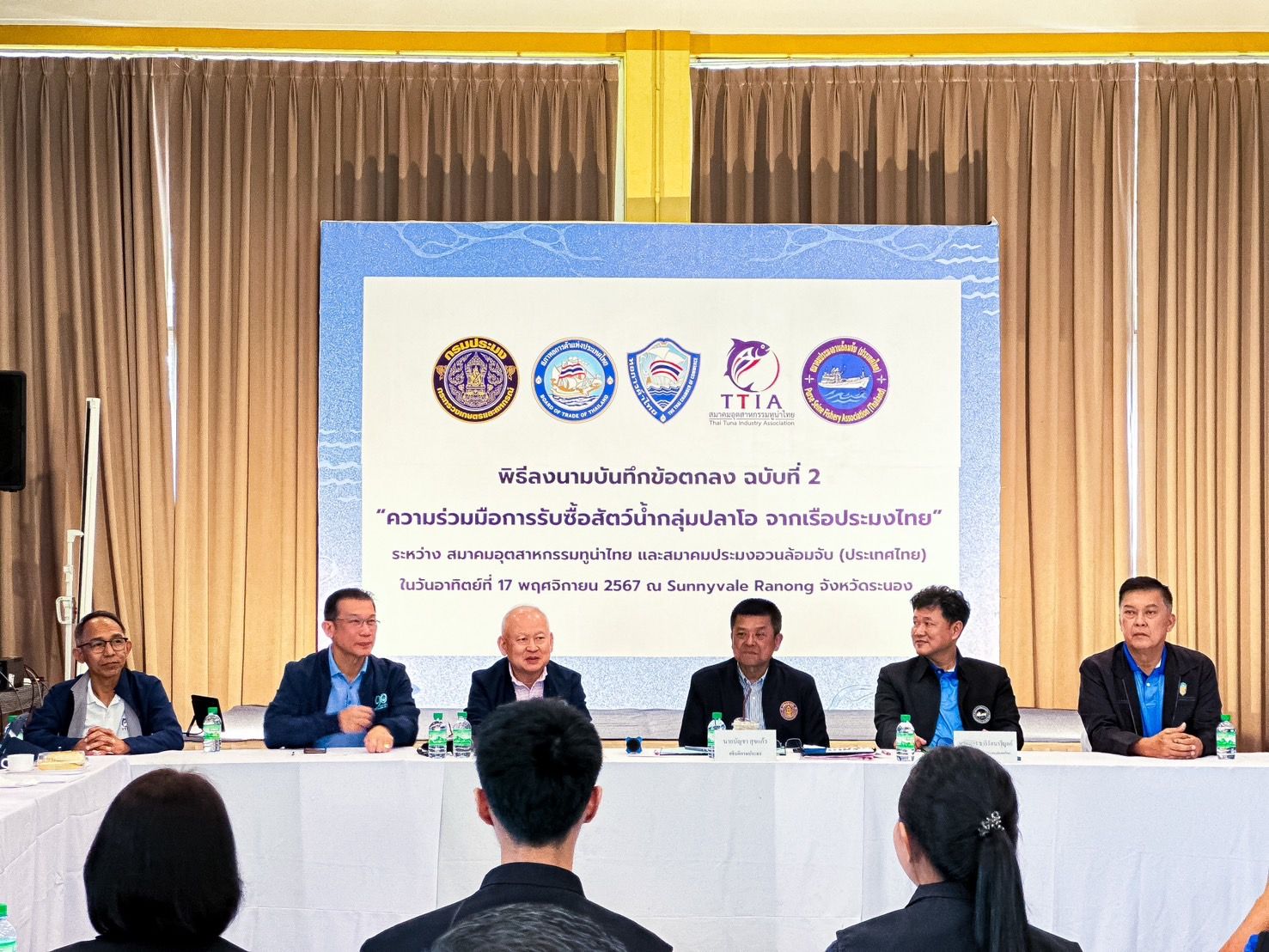 Group of men at a conference table with a large screen displaying official logos.