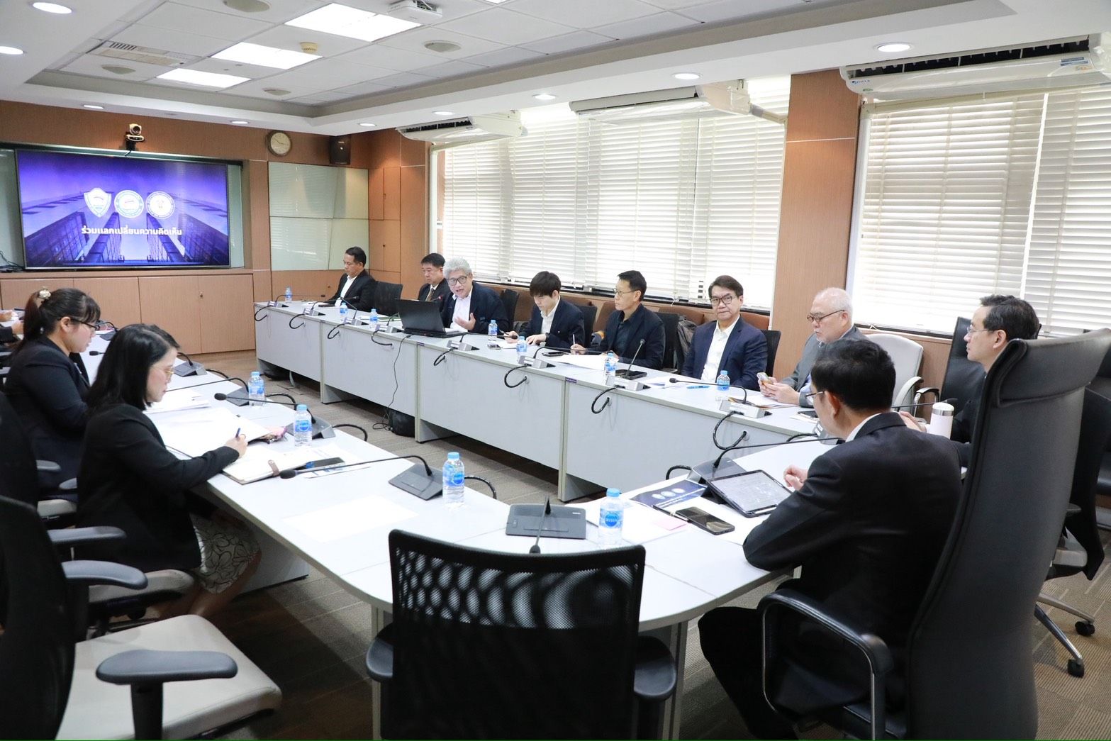 Professionals attend a meeting in a conference room with a large screen displaying graphics.