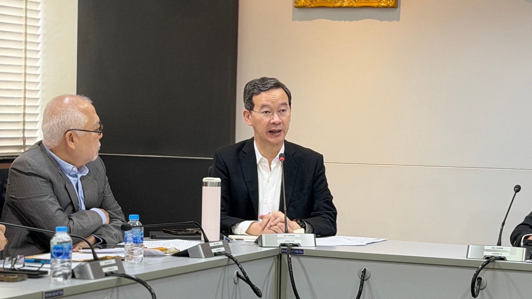 Asian man in a dark suit speaking at a conference table, another man observes.