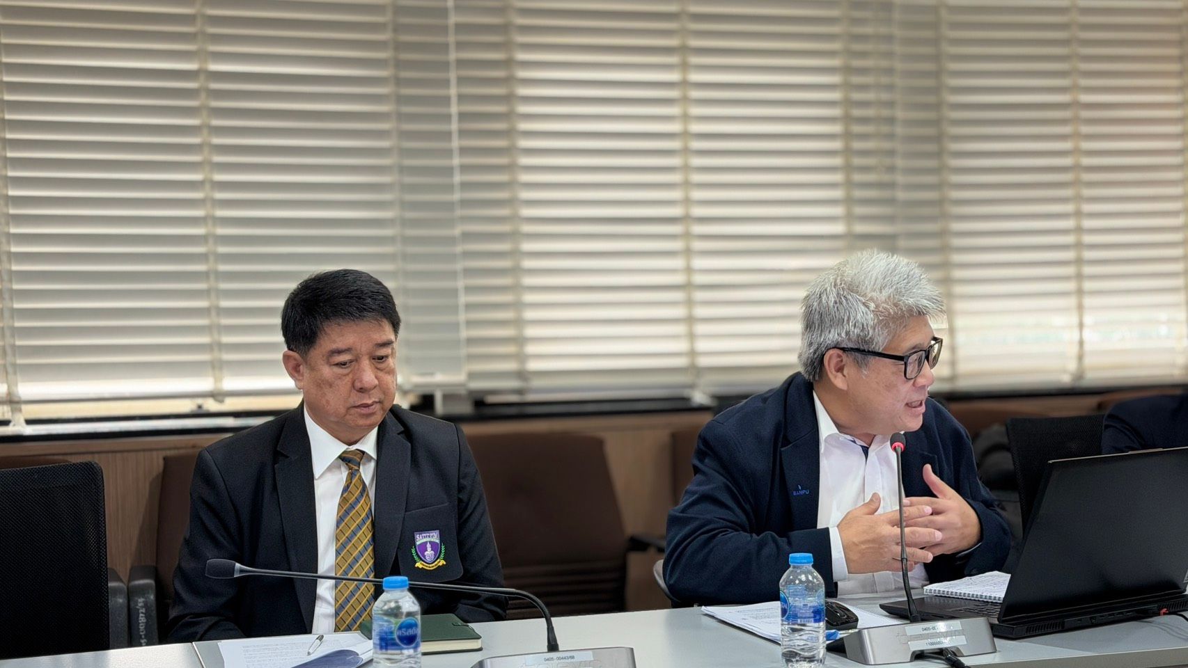 Two men, one speaking and gesturing, the other listening, at a conference table.