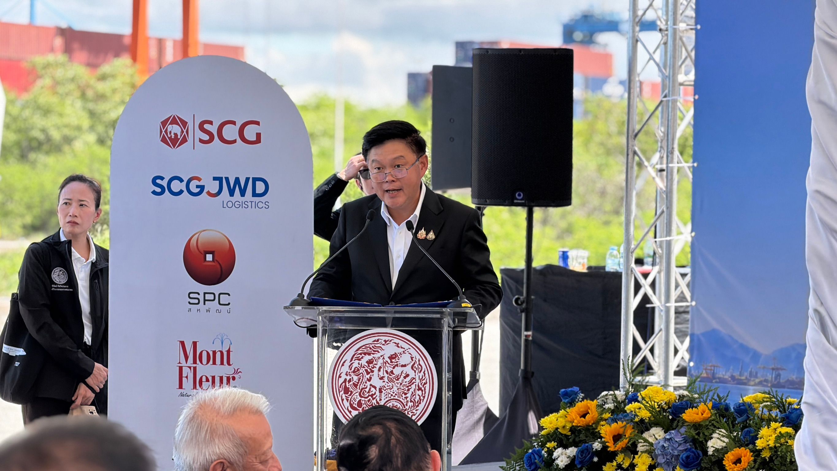 Man in suit speaking at a podium during a corporate event with SCG JWD logos.