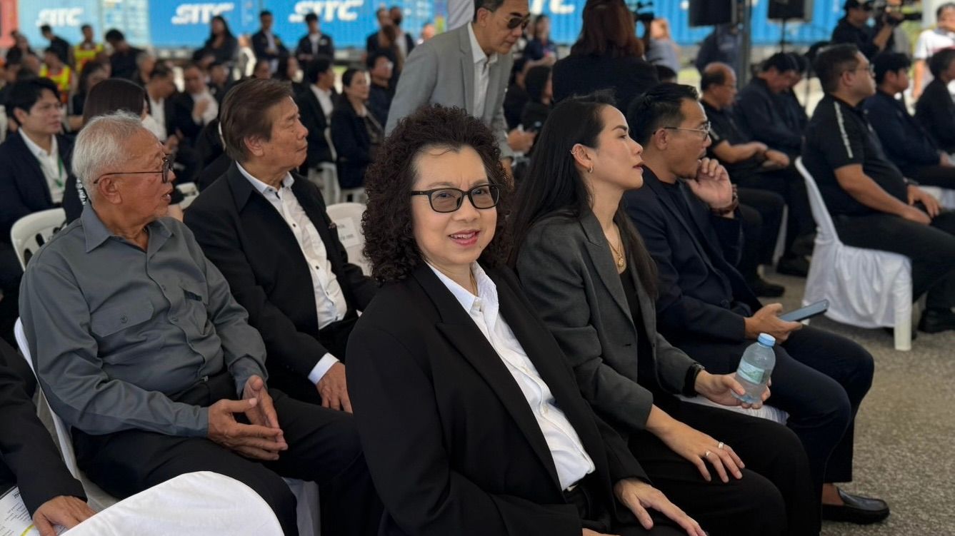 A woman in glasses and a dark suit smiles, seated among attendees at an outdoor event.